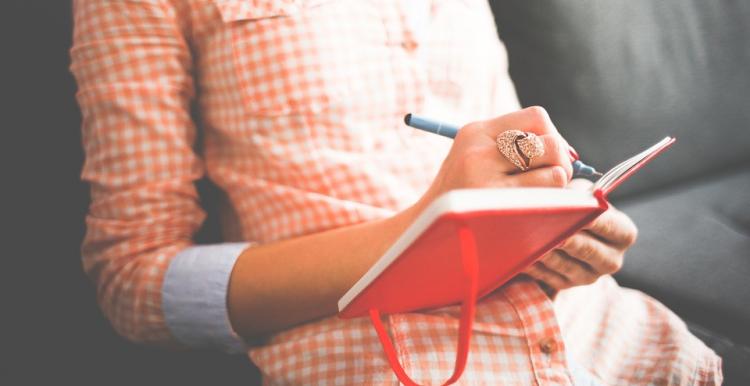 Woman in checked shirt sitting down and writing in a red notebook